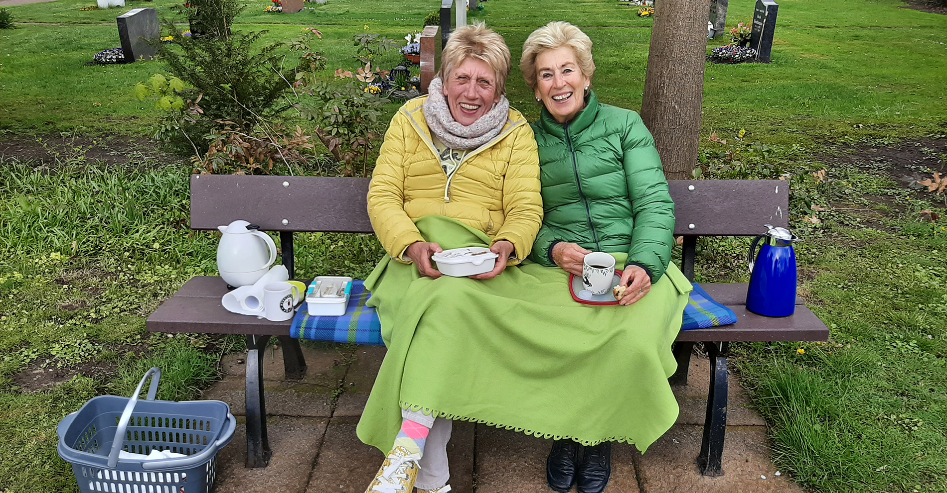 Zwei Frauen sitzen mit Kaffee auf einer Bank und unterhalten sich auf dem Friedhof